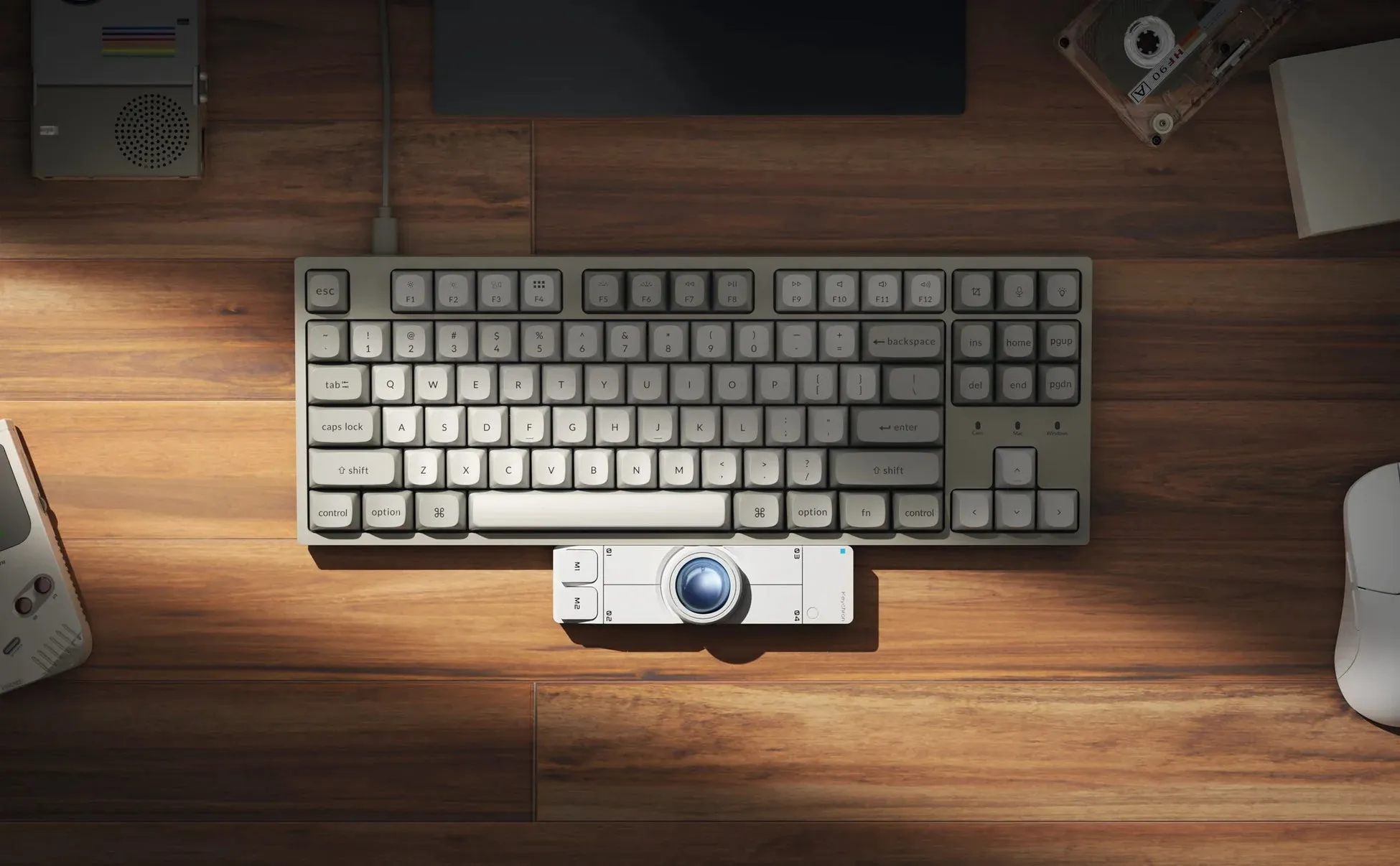 木製のテーブルの上に置かれたガジェットの俯瞰図。キーボード、トラックボール、マウス、カセットテープ、およびゲームボーイ。