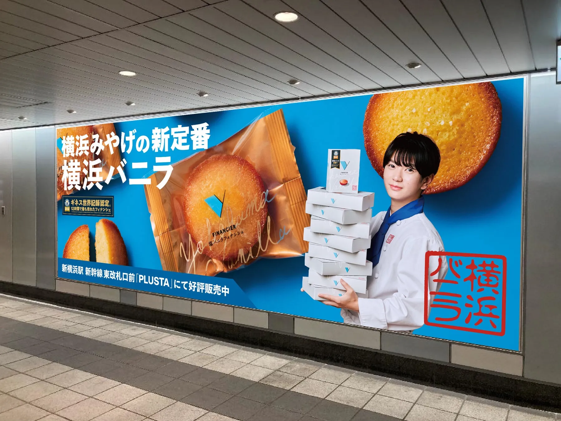 横浜みやげの新定番 横浜バニラというお菓子の広告。若い女性が商品を持ち、大きなバニラフィナンシェが背景に配置されています。広告は駅の壁面に掲示されています。