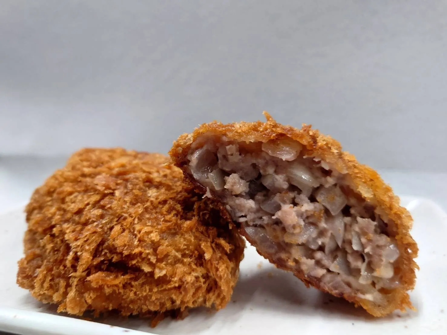 揚げ物のコロッケが2つ、手前に1つと半分に切られたものが写っている。衣はカリカリで、具材は細かく刻まれたものが見える。