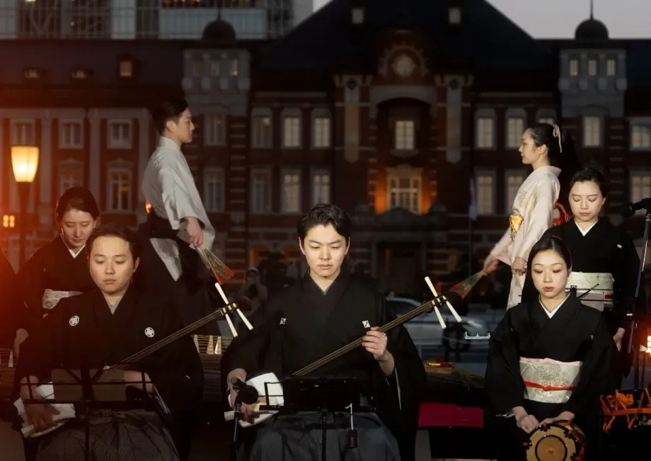東京駅を背景に、着物姿の演者たちが三味線や太鼓などの和楽器を演奏している夜の屋外パフォーマンスの様子